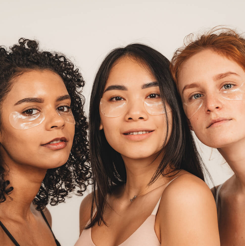 Three women of diverse ethnic backgrounds wearing under-eye gel patches, posing closely together against a neutral background, promoting skincare and self-care