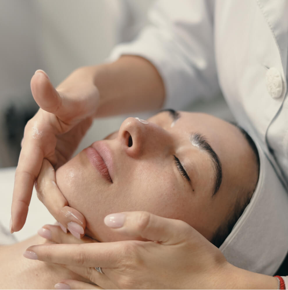 A woman lying down with her eyes closed, receiving a gentle facial massage from a skincare professional
