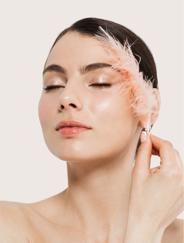 A woman with flawless, glowing skin and closed eyes gently touches her cheek with a soft, pale pink feather